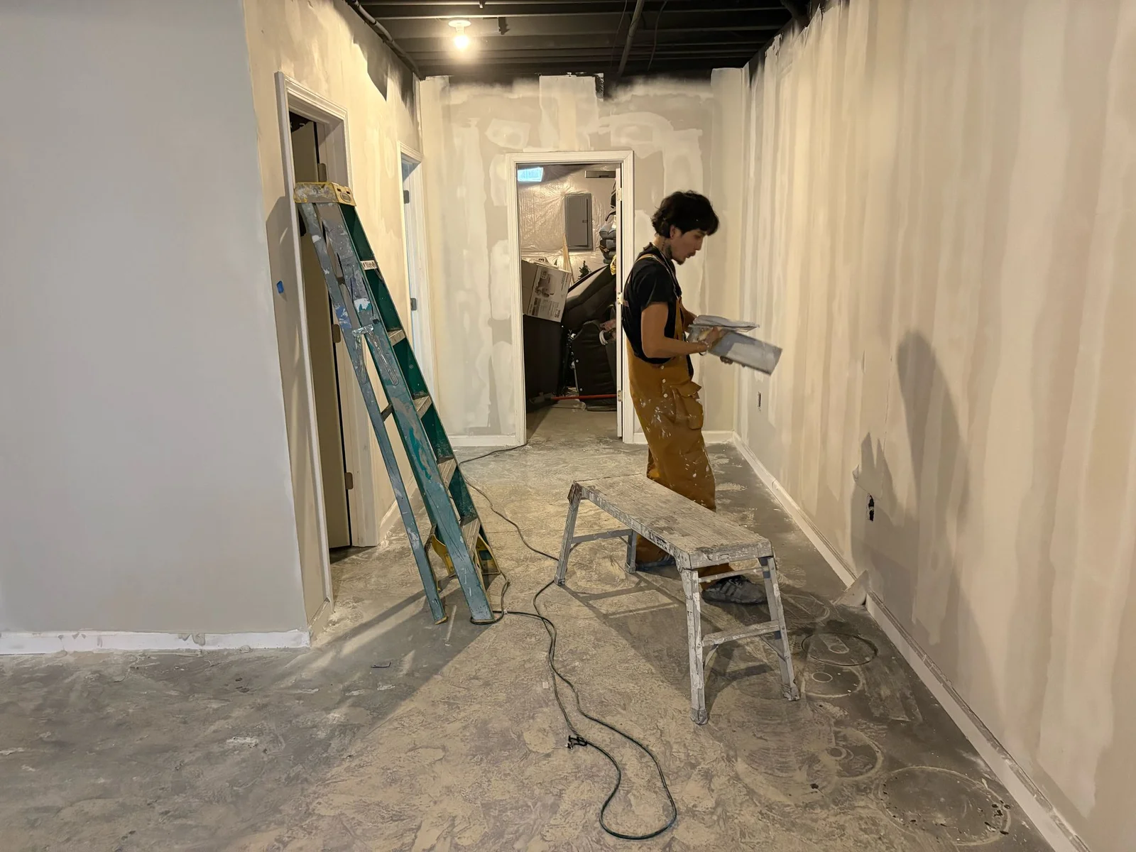 Commercial painter applying finishing touches to freshly prepared interior walls, with a ladder and equipment in a partially renovated space.