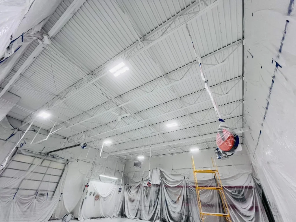 Commercial ceiling painted with dryfall technique, featuring bright lighting and protective coverings, showcasing a professional painting environment.