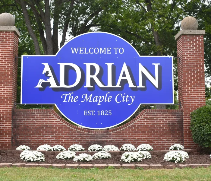 Welcome sign for Adrian, Michigan, featuring "Welcome to ADRIAN" and "The Maple City," established 1825, surrounded by white flowers and greenery.