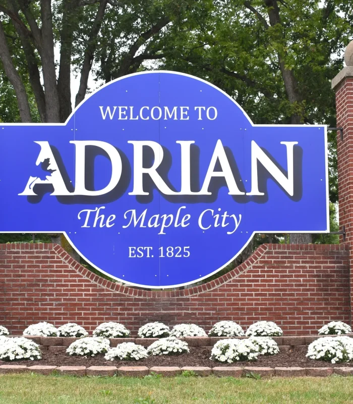 Welcome sign for Adrian, Michigan, featuring blue and white colors, stating "Welcome to ADRIAN The Maple City EST. 1825," surrounded by landscaped flowers and trees.