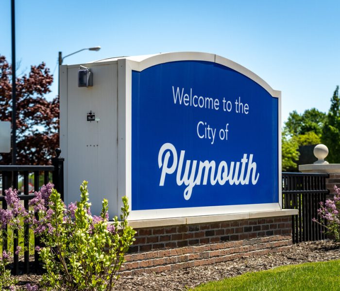 Welcome sign for the City of Plymouth, MI, featuring vibrant blue background and white lettering, surrounded by greenery and flowers, symbolizing community and local identity.